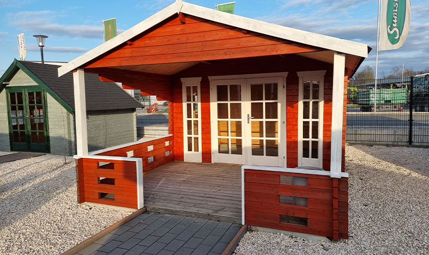 Ein rotes Holzhaus mit weißen Fensterrahmen, Vordach und heller Terrasse auf Kiesfläche.