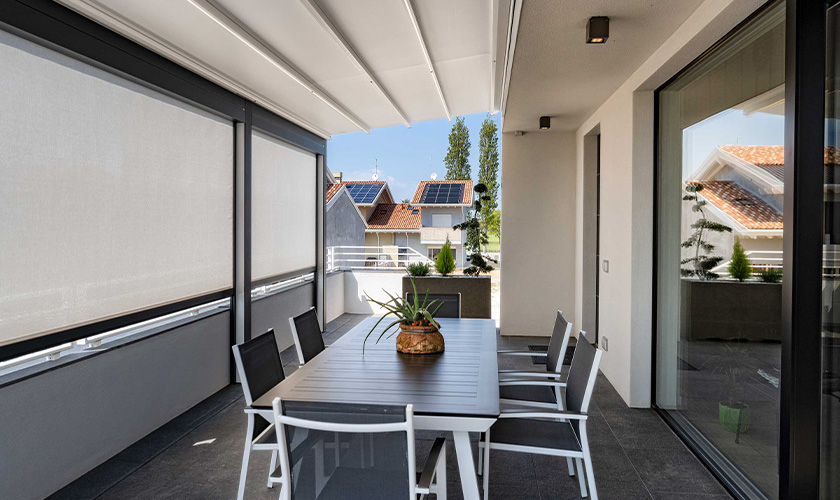 Überdachte Terrasse mit Esstisch, grauem Boden, Pflanzen und seitlichen Sichtschutzrollos.