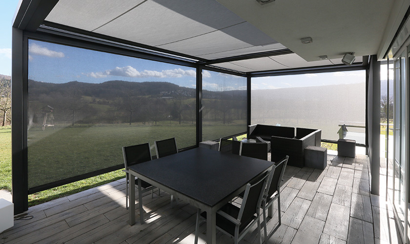 Moderne Terrasse mit Pergola, grauen Möbeln und Sichtschutz; Blick auf Natur und Hügel im Hintergrund.