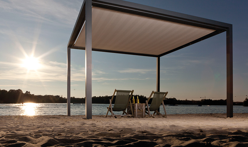 Zwei leere Liegestühle unter einer modernen Pergola am Strand bei Sonnenuntergang.