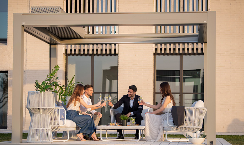 Vier Personen stoßen unter einer modernen Pergola mit Wein an. Sie lächeln, helle Einrichtung.