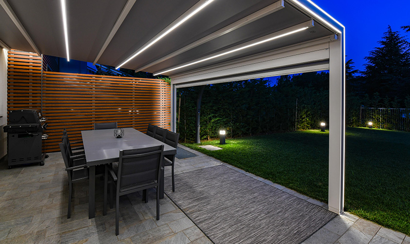 Überdachte Terrasse mit Tisch, Stühlen und Grill, beleuchtet, angrenzend an einen gepflegten Garten.