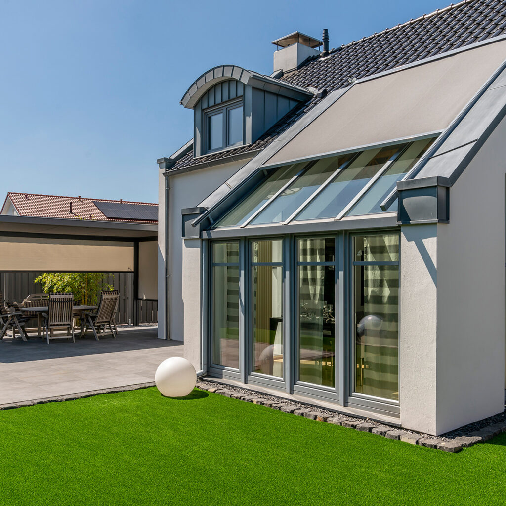Modernes Haus mit Wintergarten, großen Fenstern und gepflegtem Rasen unter klarem, blauem Himmel.