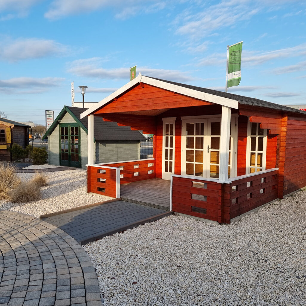 Rotes Gartenhaus mit Veranda, daneben ein grünes Haus. Blauer Himmel, Steine im Vordergrund.