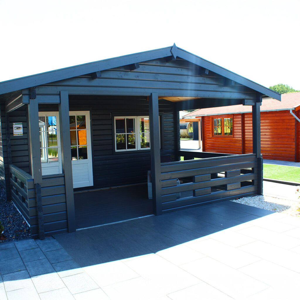 Dunkelgraues Holzhaus mit Veranda und Fenstern, sonnig beleuchtet, daneben ein rotes Holzhaus.