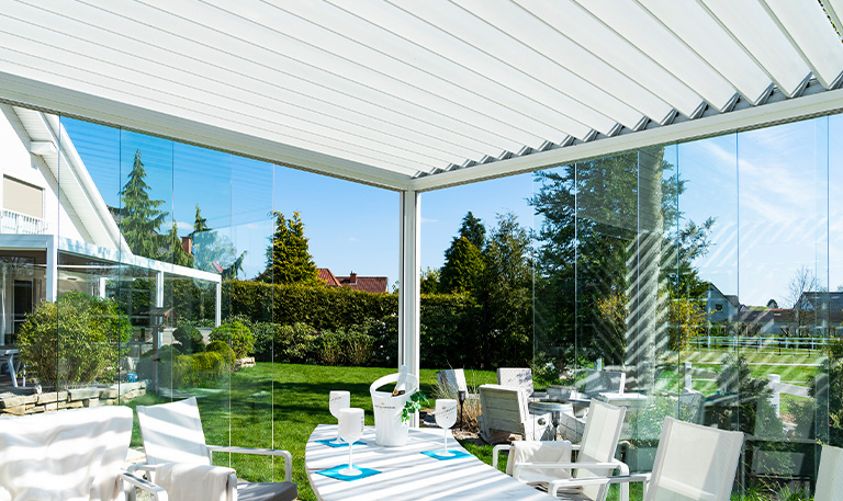Eine moderne, helle Terrasse mit Glasschiebewänden, weißen Möbeln und einem gepflegten Garten.