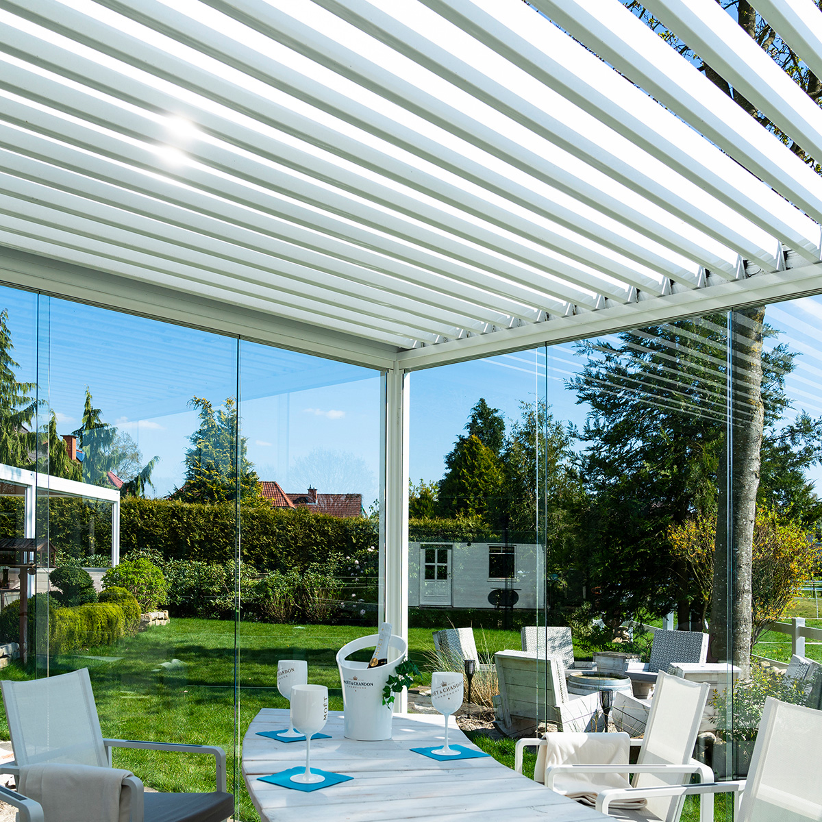 Moderne Terrasse mit Glaswänden und grüner Aussicht, weiße Möbel, schattenspendende Lamellen.