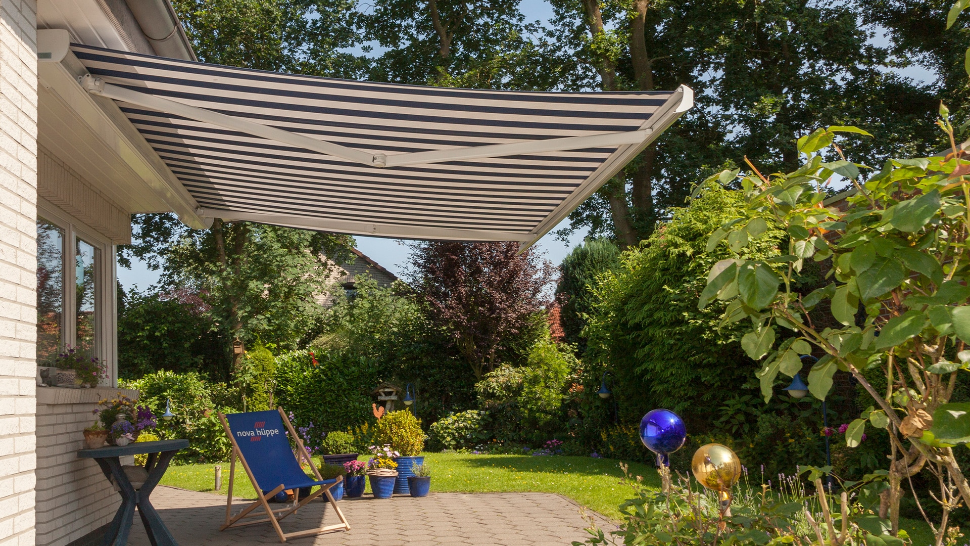 Markise im Garten, gestreift, blau-weiß, blühende Pflanzen, zwei Stühle, sonniger Tag.