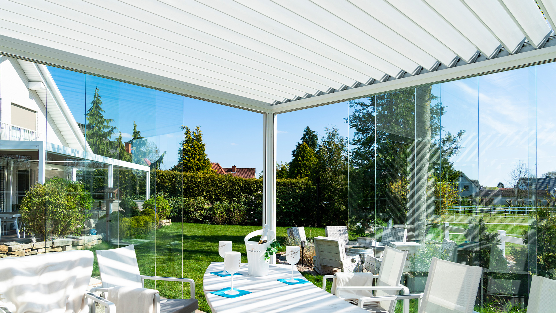 Eine sonnige Terrasse mit weißen Möbeln und verglasten Wänden, mit Blick auf den Garten.