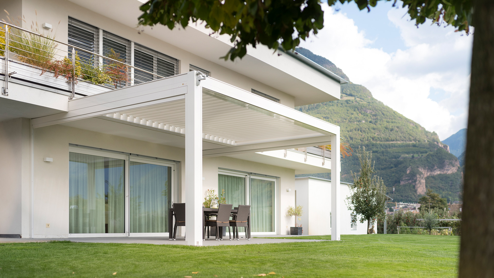 Moderne Terrassenüberdachung an einem weißen Haus mit Bergkulisse und grünem Rasen.
