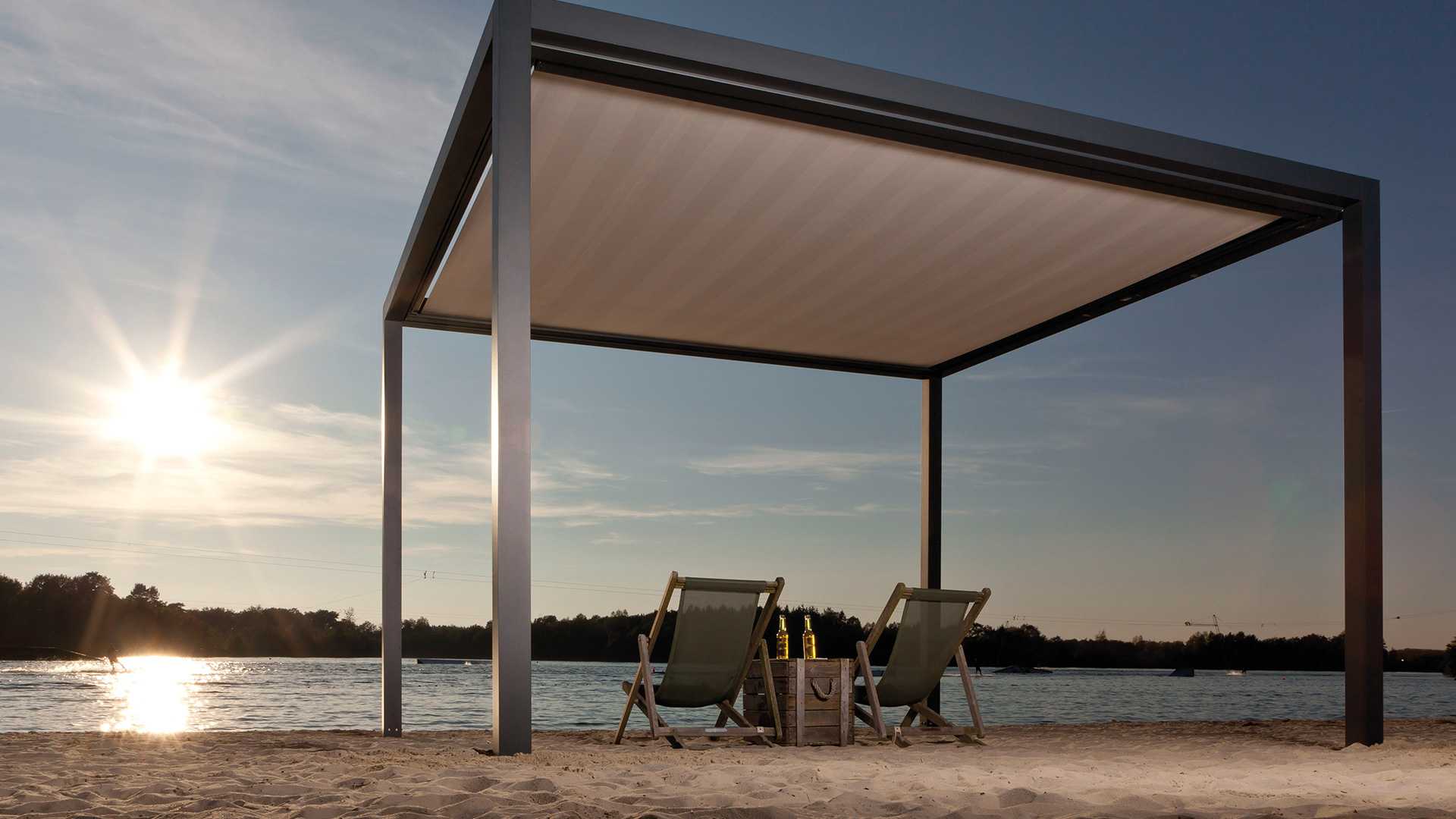 Zwei leere Strandliegen unter einem modernen, offenen Pavillon bei Sonnenuntergang am Seeufer.