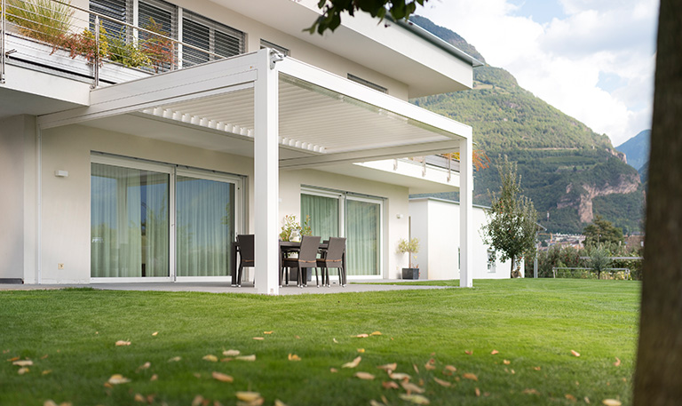 Modernes weißes Gebäude mit Terrasse, Rasen und Berg im Hintergrund bei bewölktem Himmel.