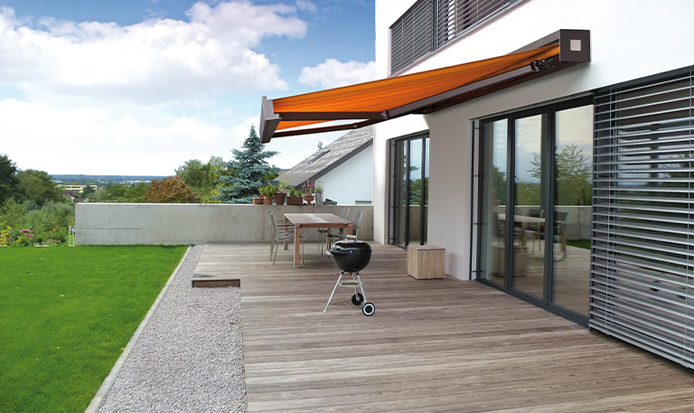 Eine Terrasse mit Grill, Holztisch, orangefarbener Markise, und Blick auf einen Rasen und Bäume.