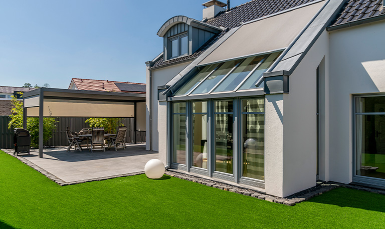 Modernes Haus mit Terrasse, großem Fenster, weißer Wand und grünem Rasen, sonniger Tag.