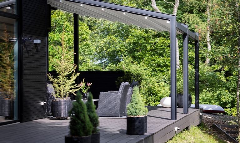 Überdachte Terrasse mit schwarzen Möbeln; grüne Bäume; moderne, geradlinige Architektur.