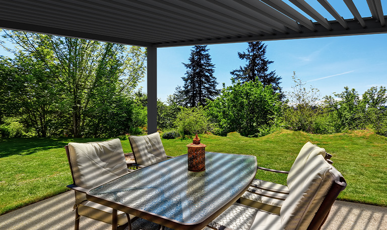 Terrasse mit Glastisch und sechs Stühlen, sonnig, grüner Gartenhintergrund, schattenspendende Pergola.